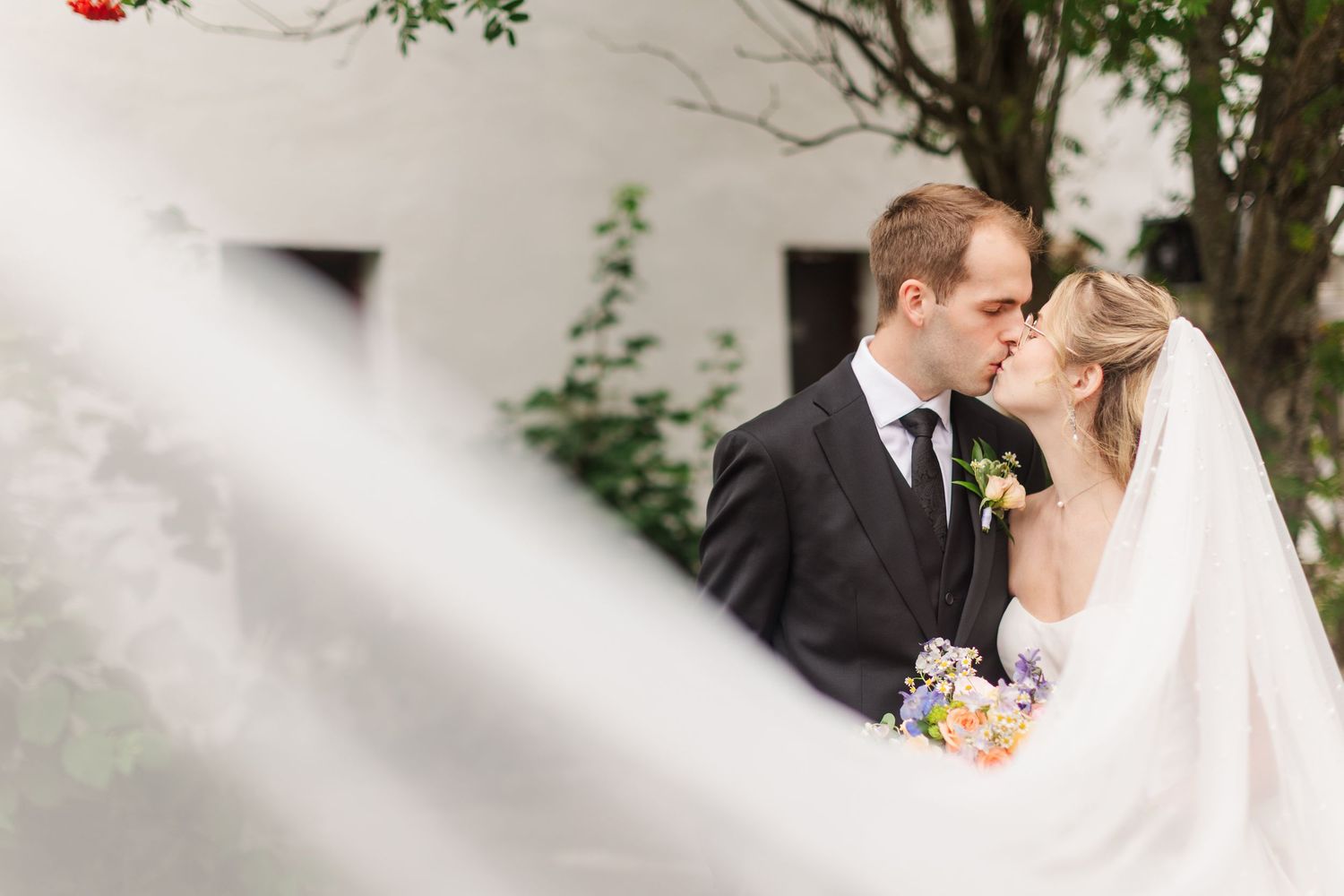 A bride and groom share a romantic kiss outdoors, with the bride's veil flowing in the foreground on their wedding day.