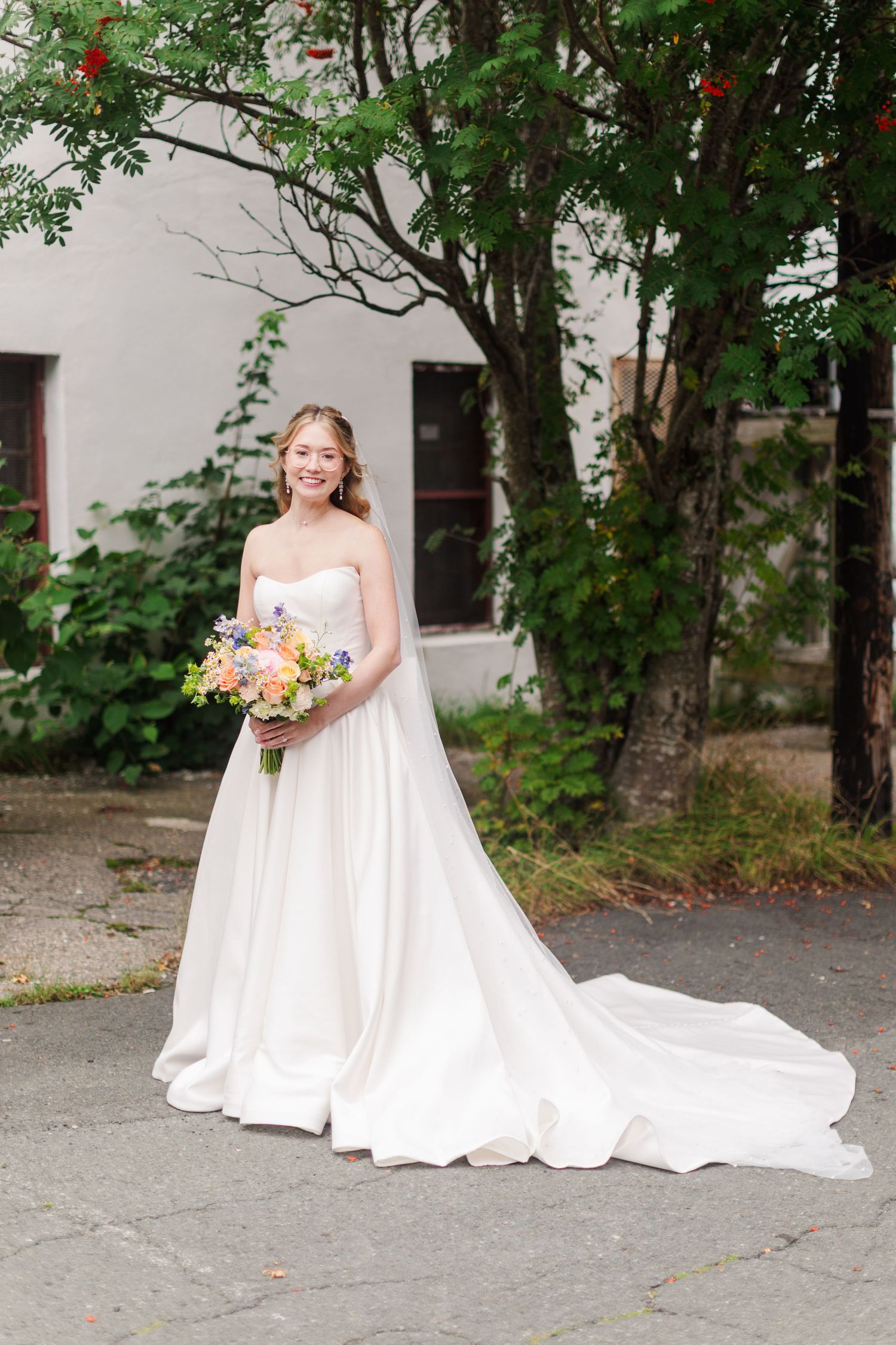 Smiling bride in a strapless white ball gown with train holds colorful bouquet outdoors near green trees.