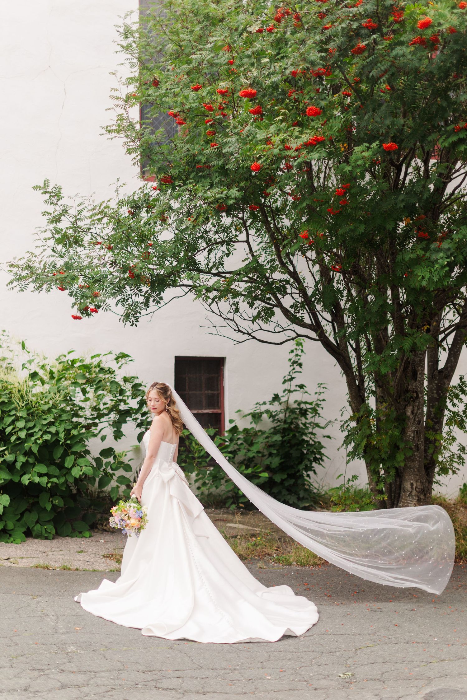 Bride in white gown with flowing veil stands near red berry tree outside white building holding colorful bouquet.
