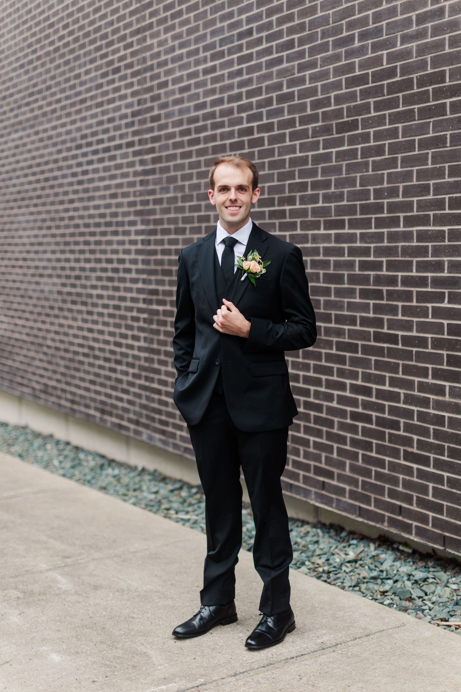 Smiling groom in black suit with boutonniere stands confidently against a brick wall on his wedding day.