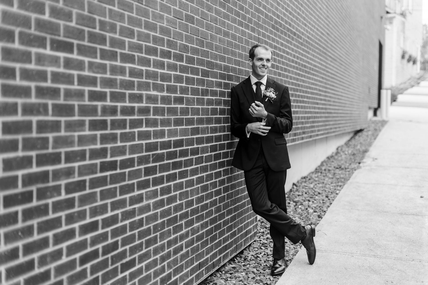 Groom in black suit leaning against brick wall, black and white wedding portrait photo.