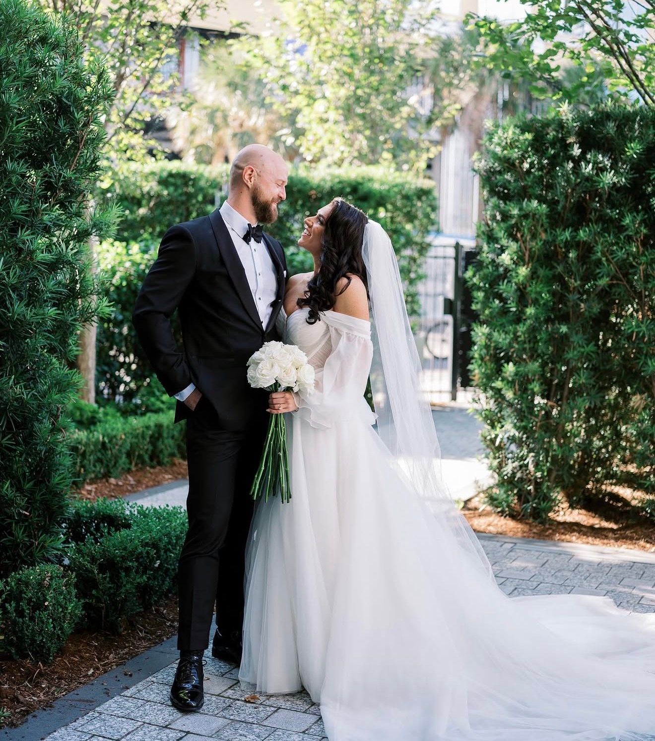 Couple sharing a romantic moment in a lush garden, groom in tuxedo and bride in off-shoulder wedding gown.