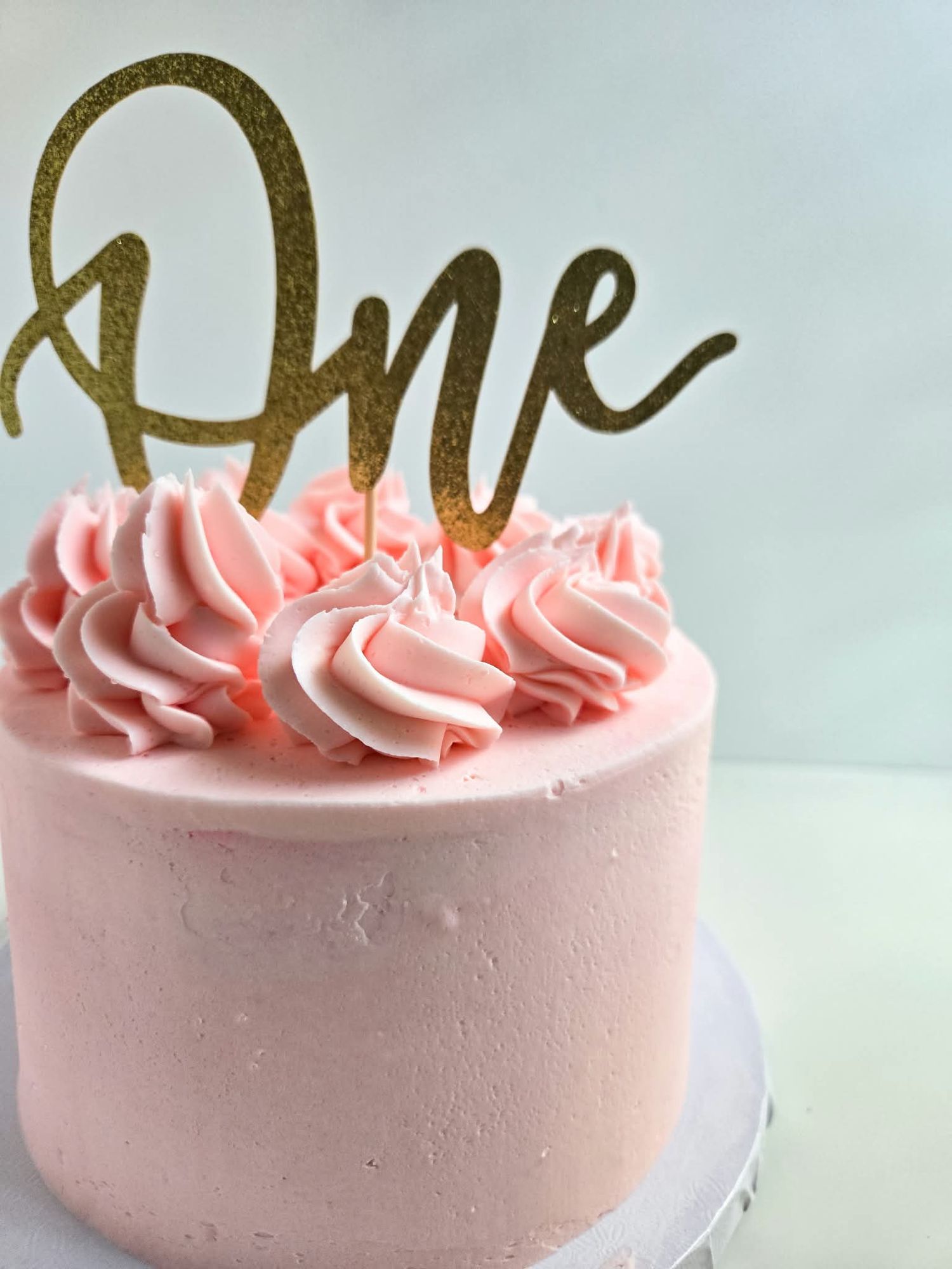 Pink first birthday smash cake with gold glitter 'One' topper and pink rosette frosting swirls.