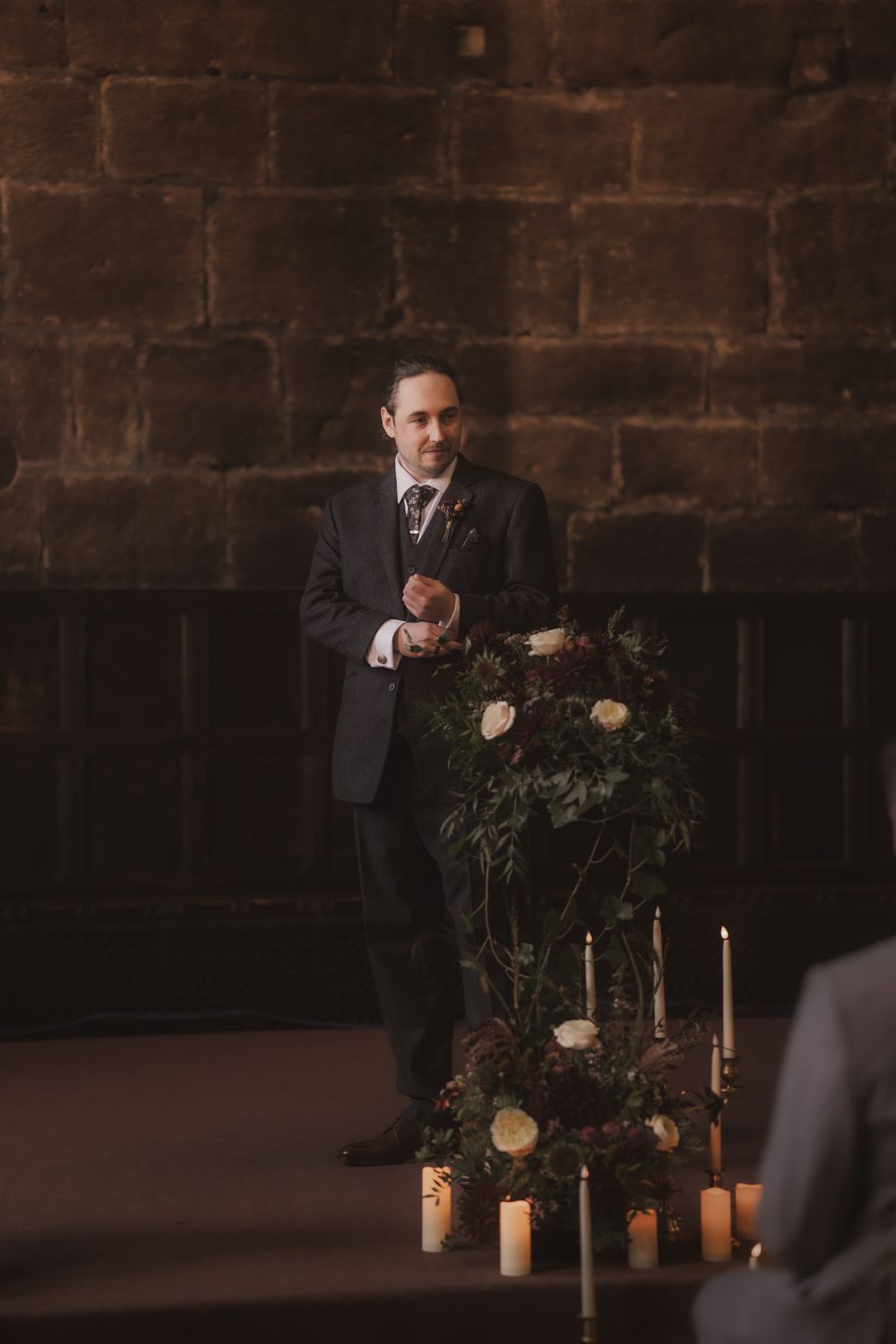 A groom in a dark suit stands beside floral arrangements and candles in a stone-walled venue during a wedding ceremony.