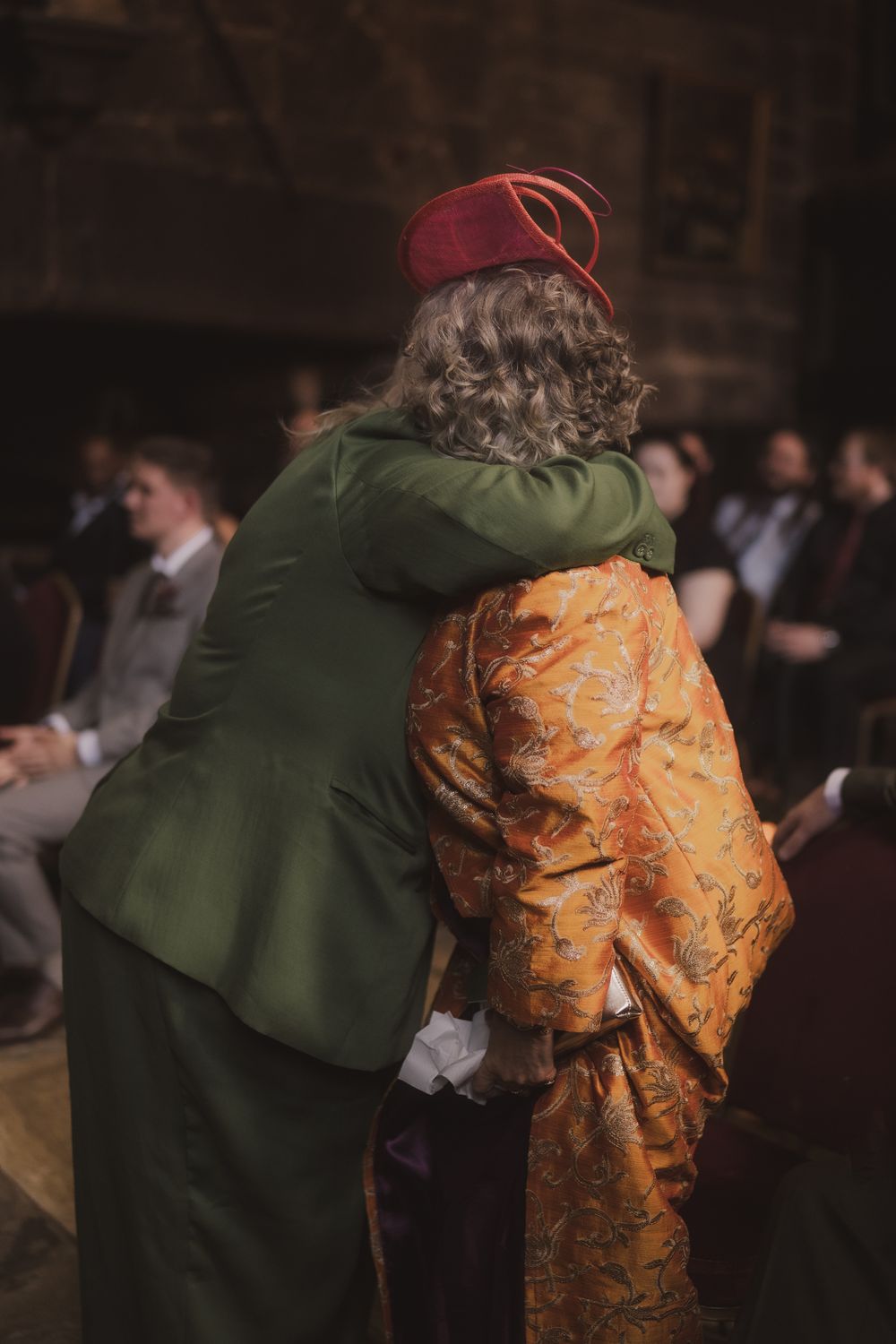 Two people in colorful vintage costumes embracing at an indoor event, one wearing a red fez hat and orange brocade robe.