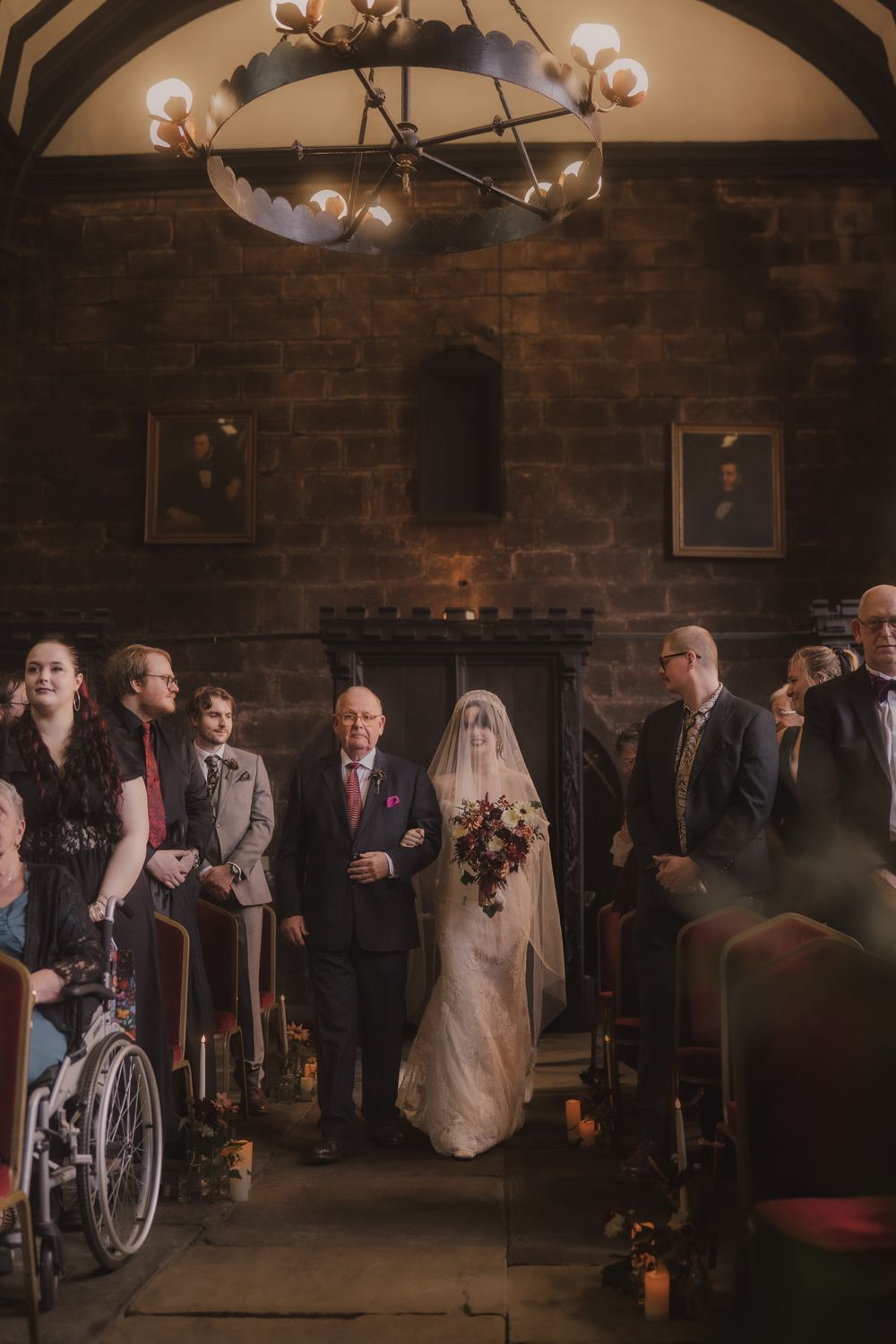 Bride walks down the aisle with her father in a rustic stone chapel, wearing a lace gown and veil, holding a burgundy bouquet.