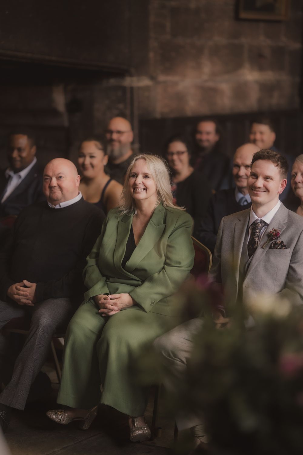 Smiling wedding guests seated in a rustic venue, featuring a woman in a green suit flanked by two men in formal attire.