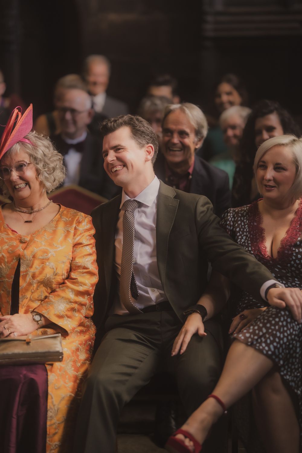 Guests laughing and smiling during a wedding ceremony, woman in orange dress with pink fascinator seated beside man in suit.