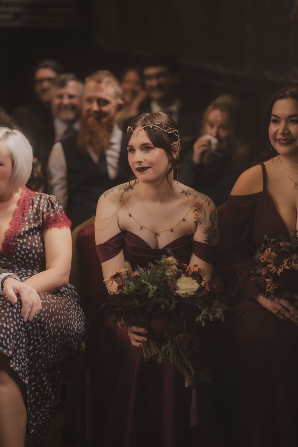 A bridesmaid in a burgundy off-shoulder gown holds a floral bouquet while standing among seated wedding guests.
