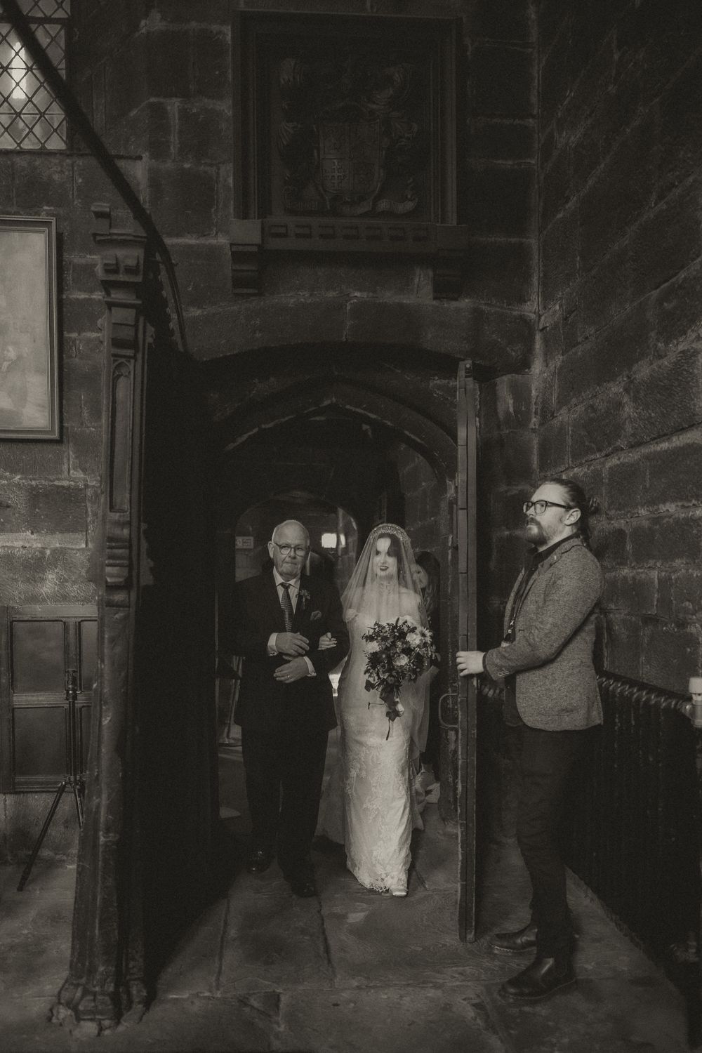 Black and white photo of a bride walking down a stone church aisle with her father and a man holding the door.