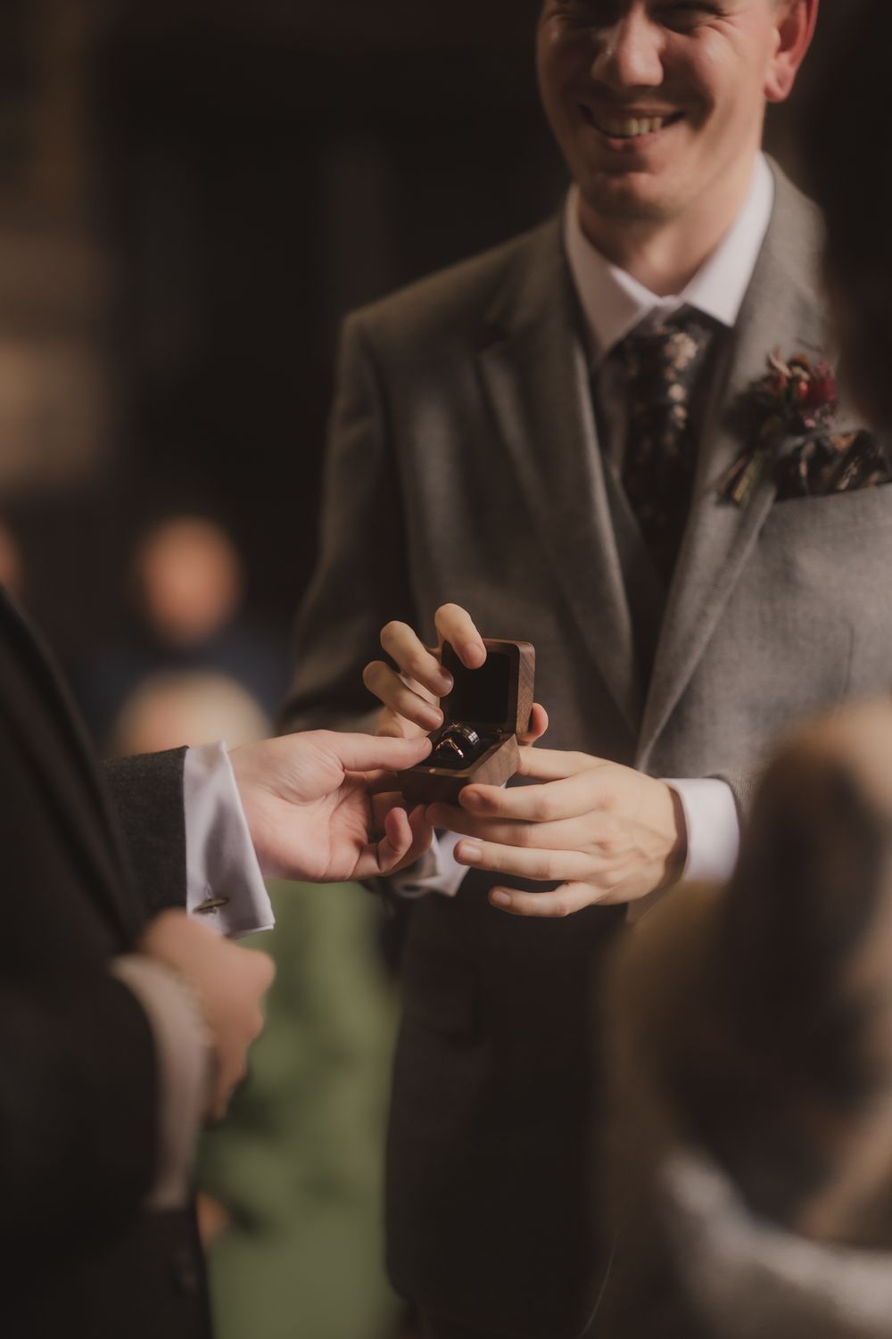 Smiling groom in gray suit holding open ring box during wedding ceremony exchange.