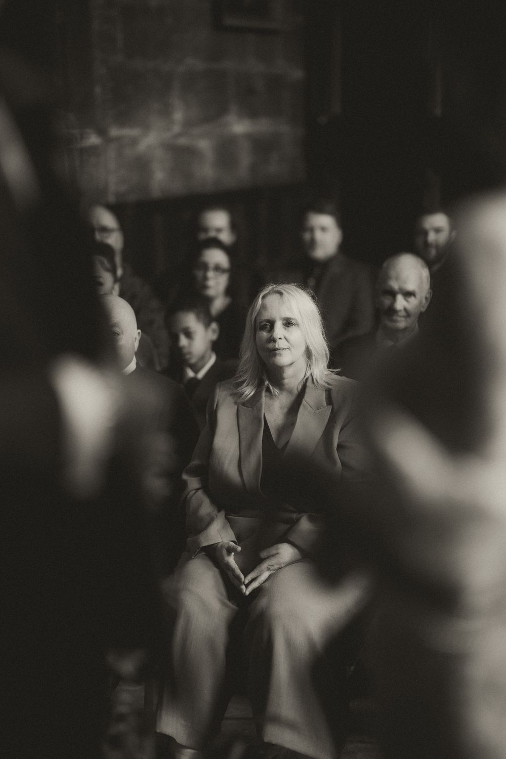 Black and white photo of a blonde woman seated in a crowd, looking forward with a calm expression.