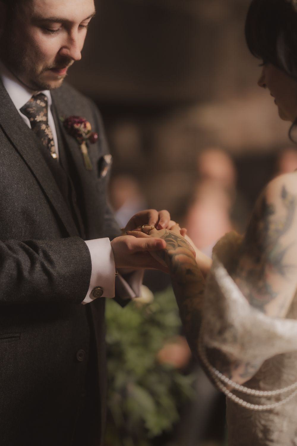 Groom in grey suit placing wedding ring on tattooed bride's finger during intimate ceremony.