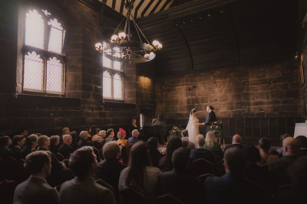 A wedding ceremony in a grand medieval stone hall with gothic windows, chandelier lighting, and guests seated before the couple.