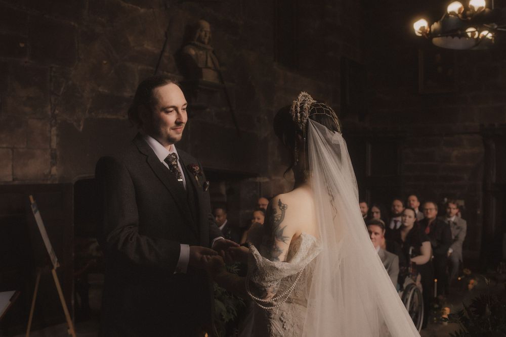 Groom reads vows to bride in elegant veil during gothic castle wedding ceremony with guests seated behind.