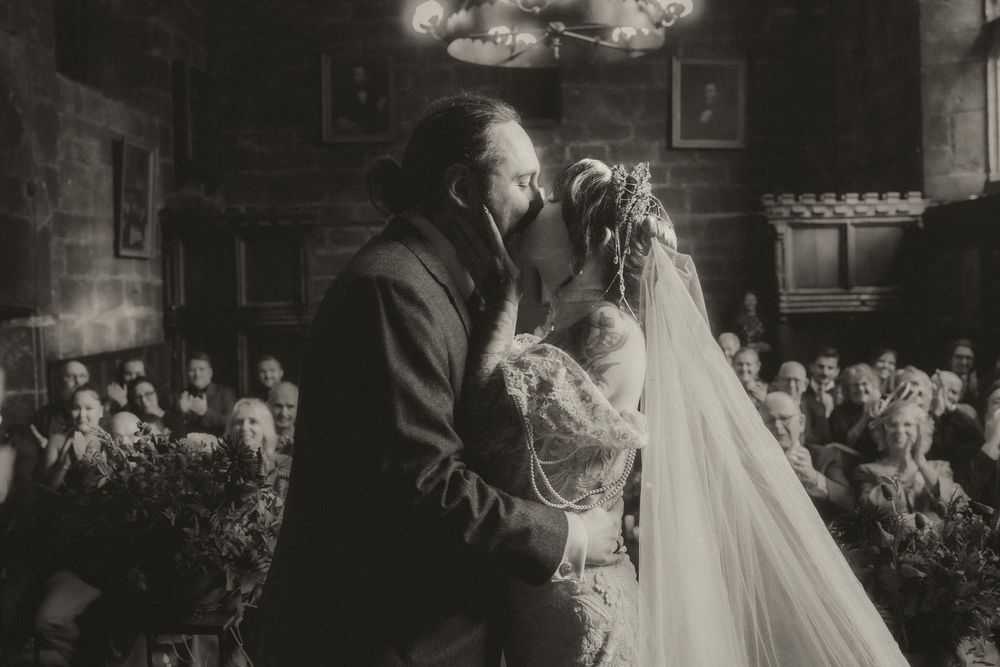 Black and white wedding photo of couple sharing first kiss at altar with guests applauding in elegant venue.