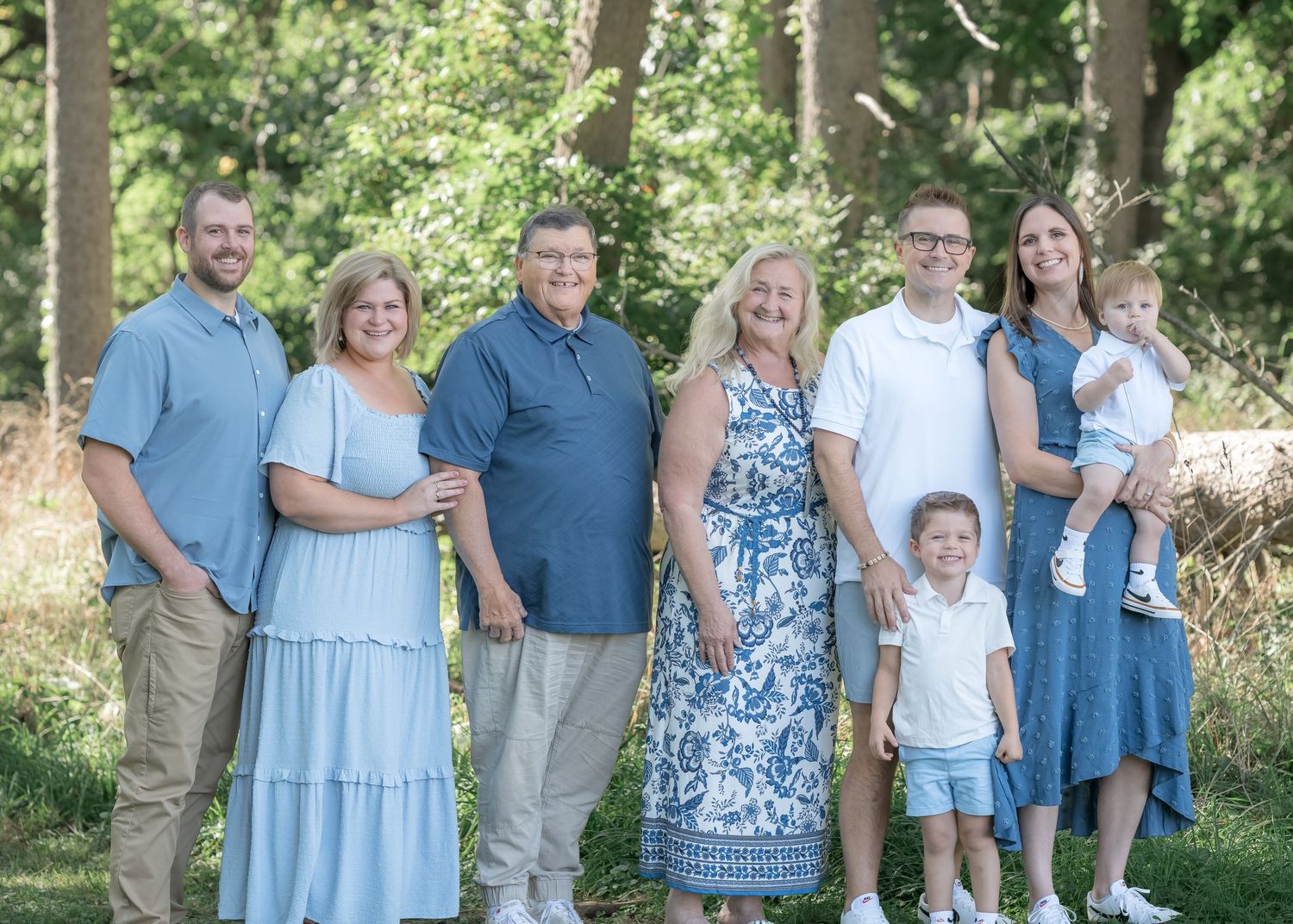 A multigenerational family of seven poses outdoors in coordinating blue outfits among lush green trees.