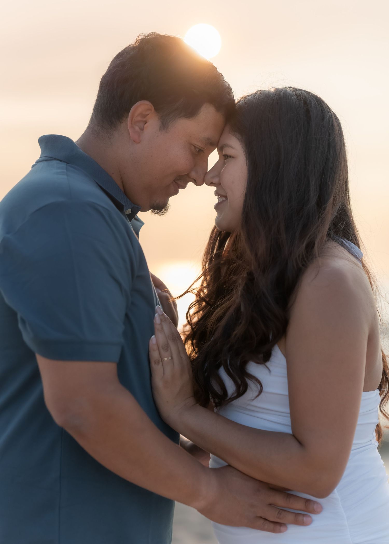 Couple sharing a romantic sunset moment on the beach, touching foreheads and smiling, golden backlight glowing behind them.