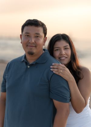 Couple posing at sunset on a beach, woman in white dress smiling with hand on mans shoulder.