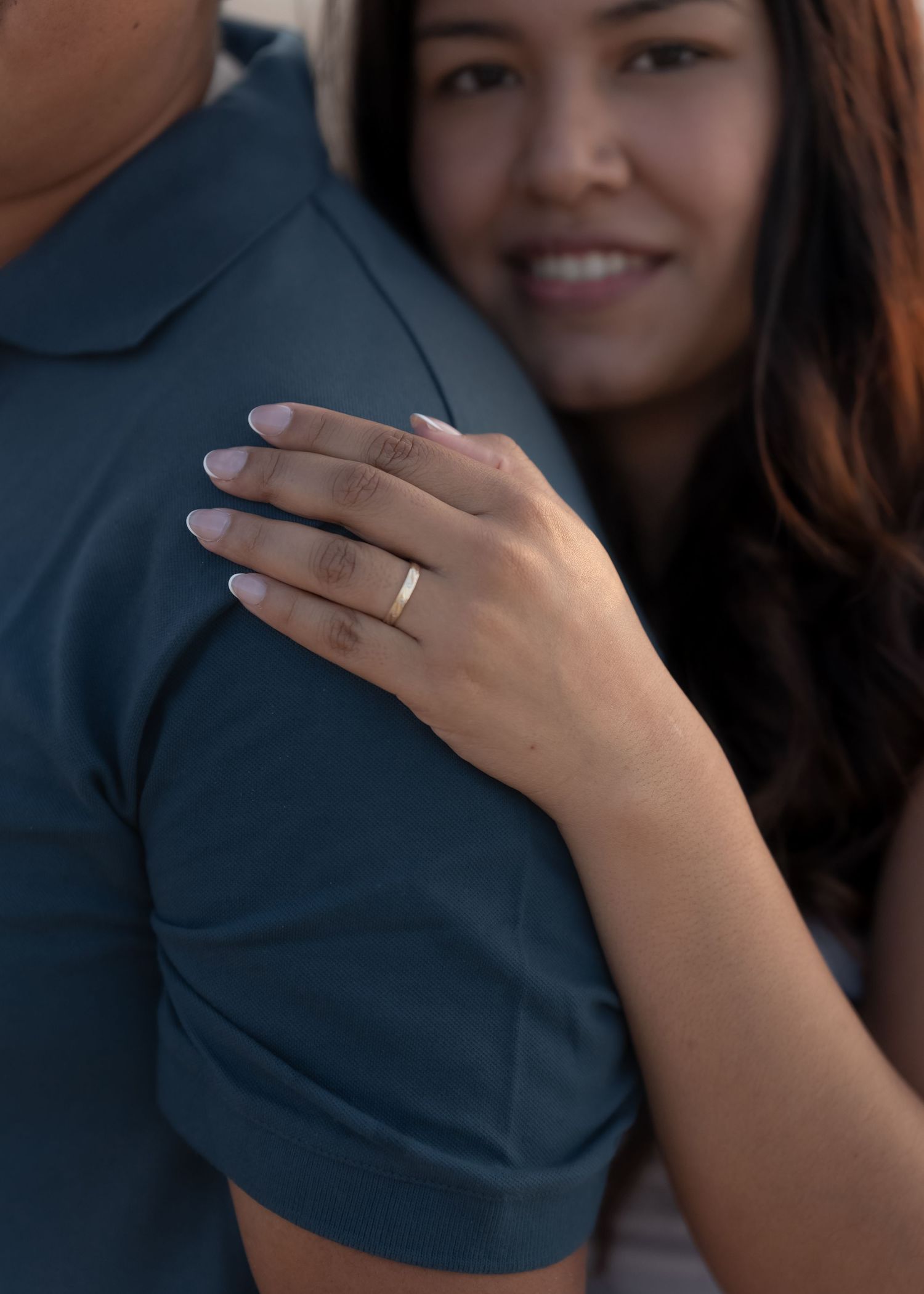 A smiling woman with a gold ring on her finger rests her hand on a man's shoulder in a romantic embrace.