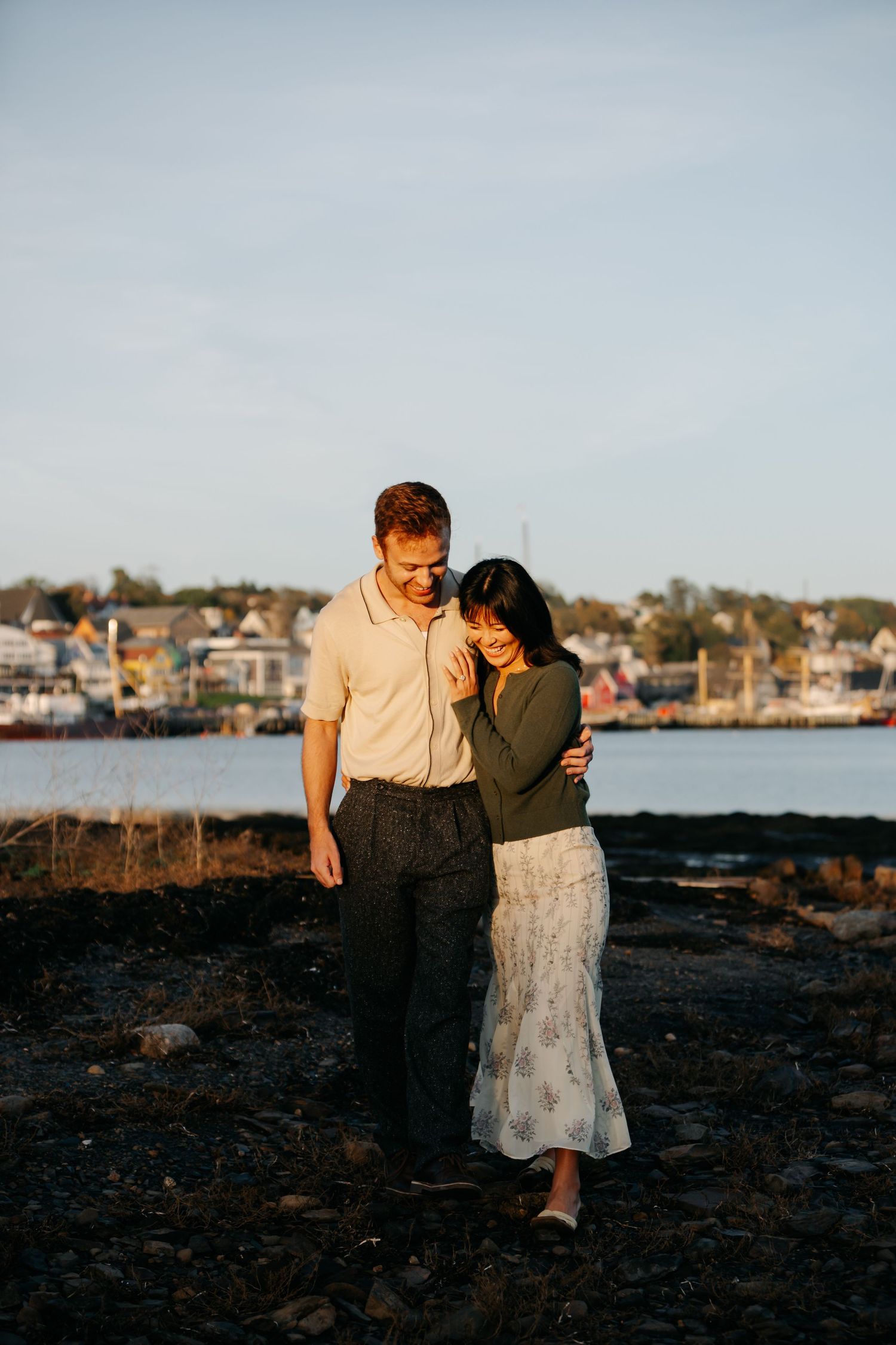 Couple embracing at golden hour by the waterfront, woman in floral skirt leaning into man in green sweater.