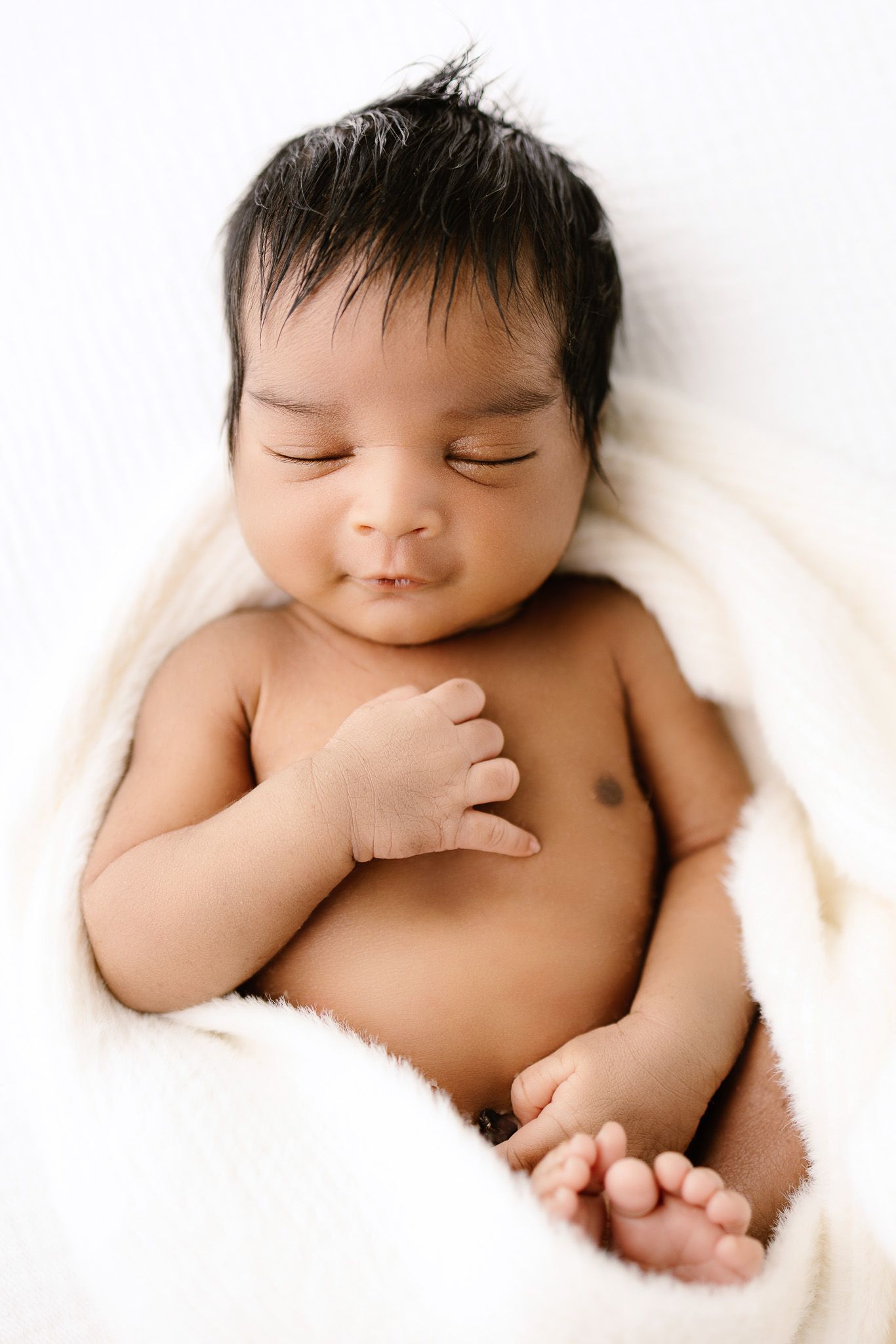 Peaceful newborn baby sleeping on white blanket, dark hair, soft skin, serene expression, professional newborn photography.