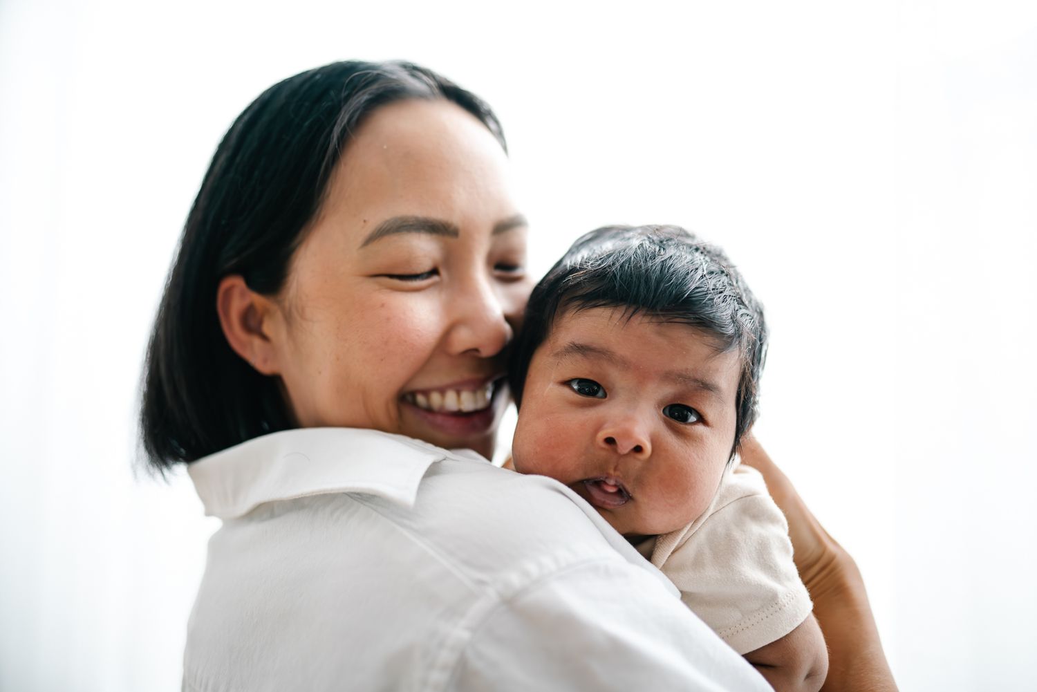 A smiling mother tenderly holds her baby close, in a loving embrace against a white background taken in-home Melbourne newborn photography.