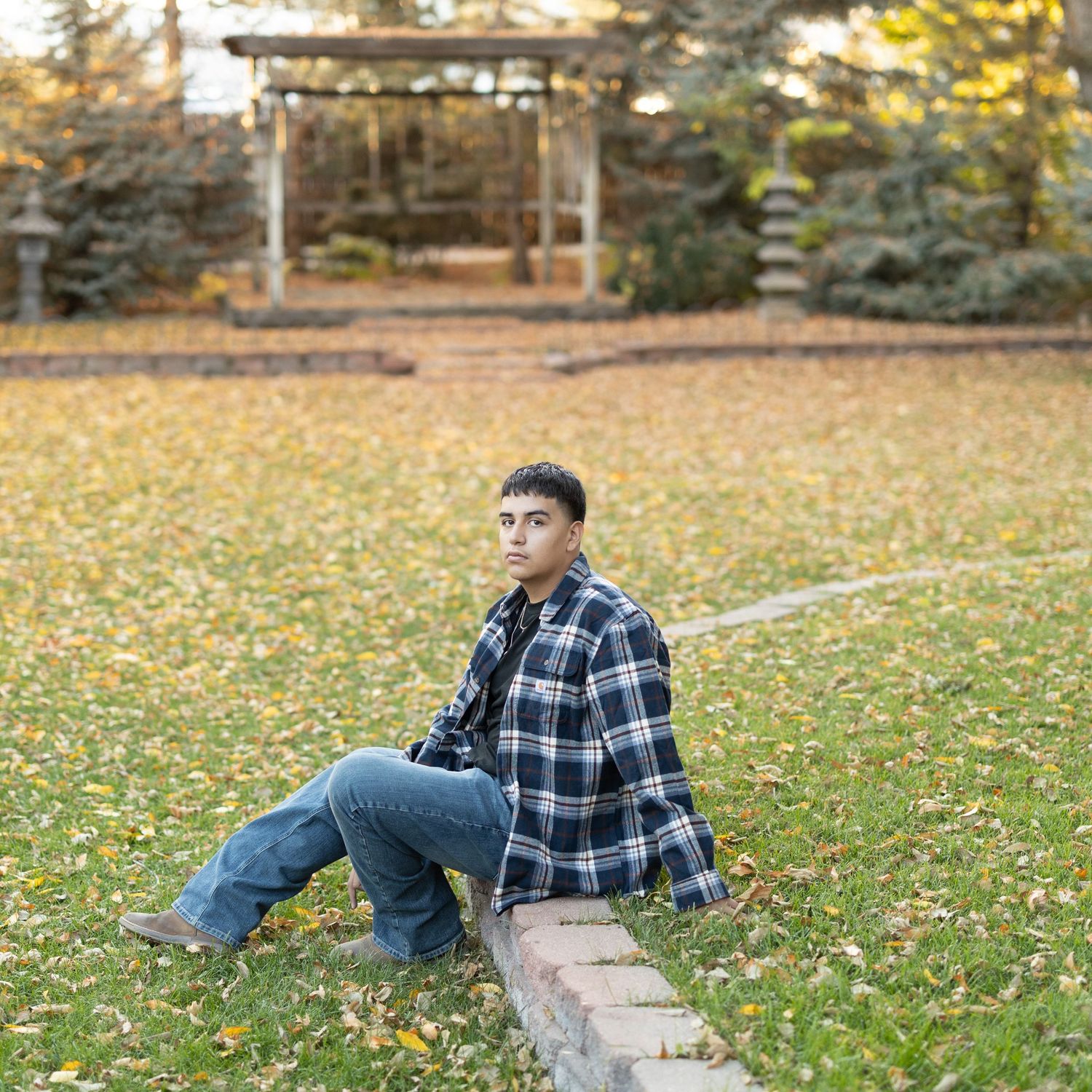 A teenage boy in a plaid flannel shirt and jeans sits on a brick border in a park with autumn leaves.