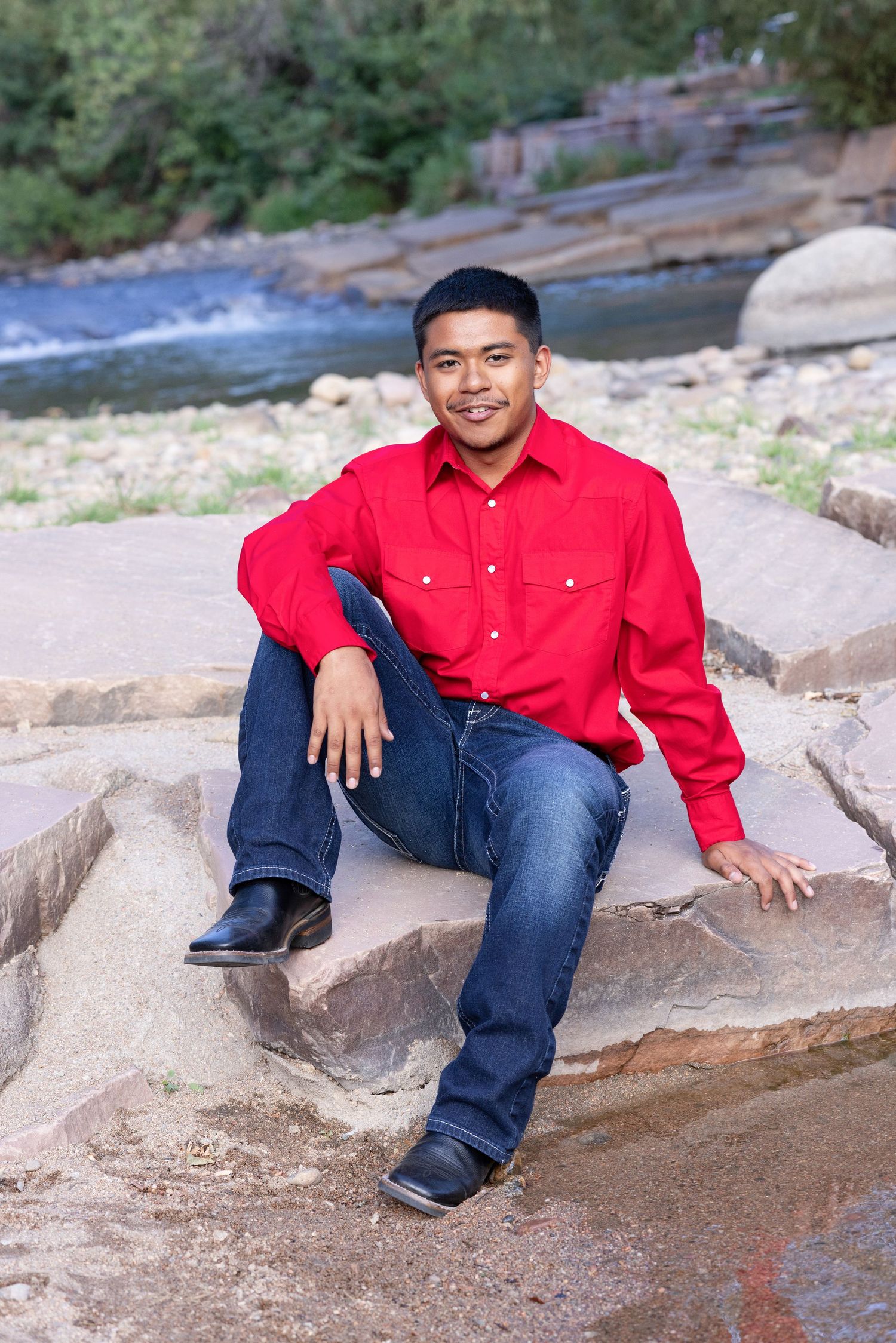 Young man in red shirt and jeans sitting on rocks by a river, smiling outdoors.