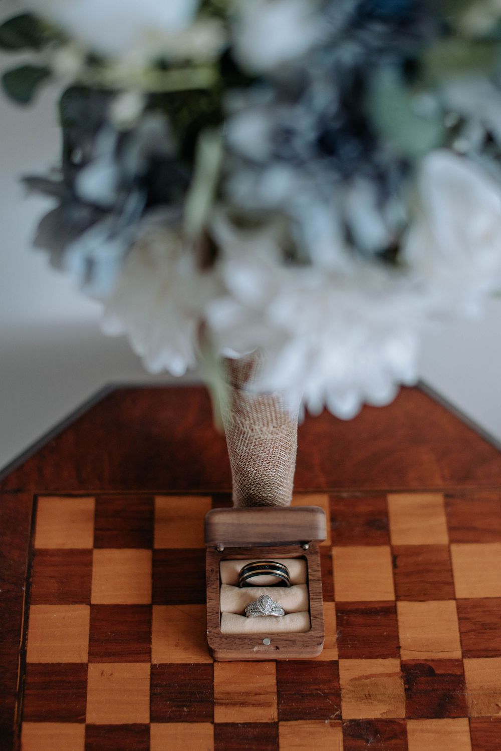 Wedding rings in a wooden box on a chessboard with a white and blue floral bouquet in the background.