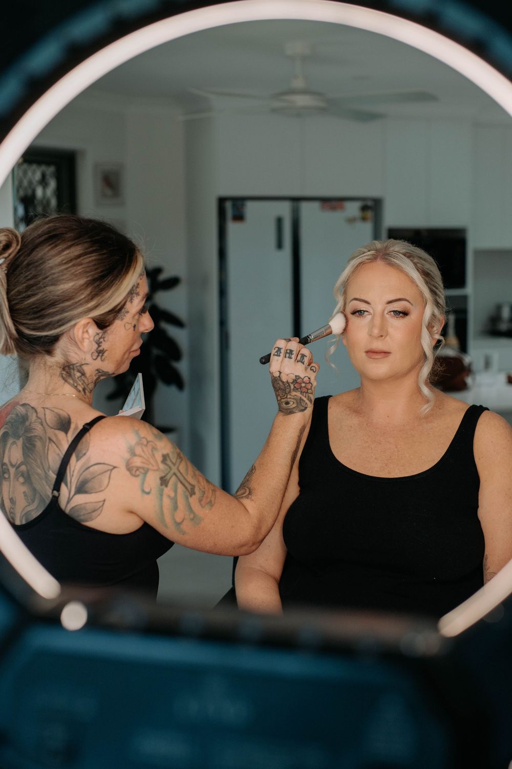 Tattooed makeup artist applying foundation to a blonde woman using a ring light mirror in a bright room.
