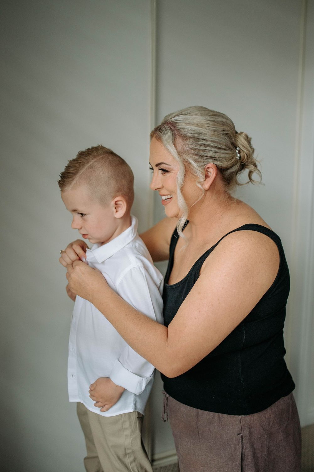 A smiling woman in a black top helps a young boy button his white dress shirt.