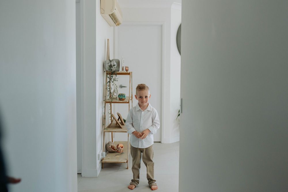 A smiling young boy in a white shirt and khaki pants stands in a bright, minimalist white hallway near a wooden shelf.
