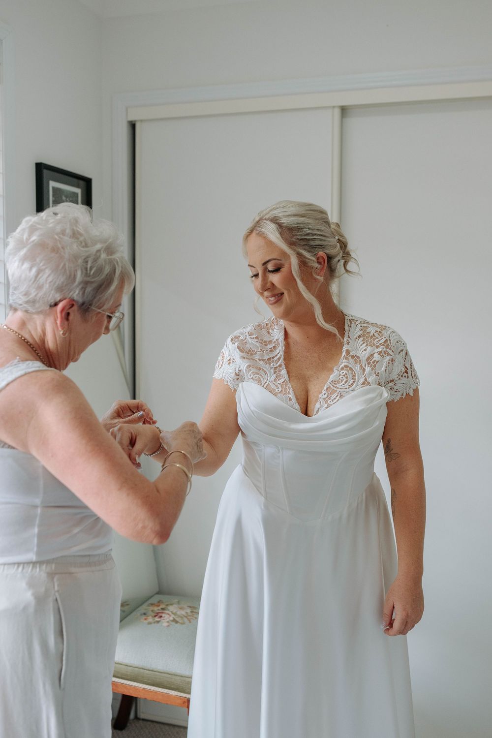 An elderly woman helps a smiling bride in a white lace wedding dress fasten her bracelet before the ceremony.