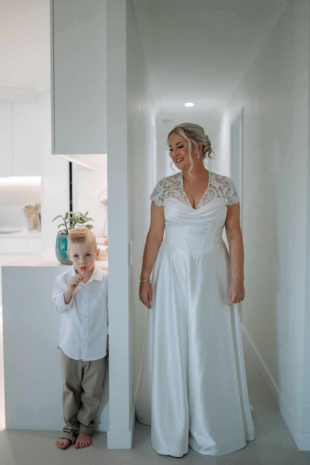 Bride in white lace wedding gown stands in hallway smiling, while young boy in white shirt peeks around corner.