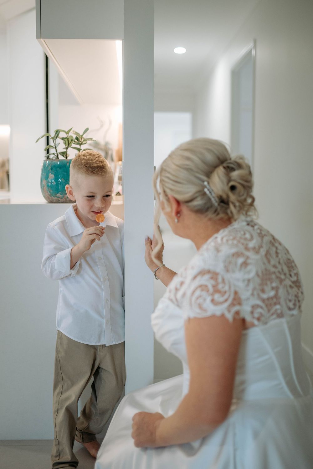A bride in a white lace dress smiles at a young boy in a white shirt and khaki pants during wedding preparations.