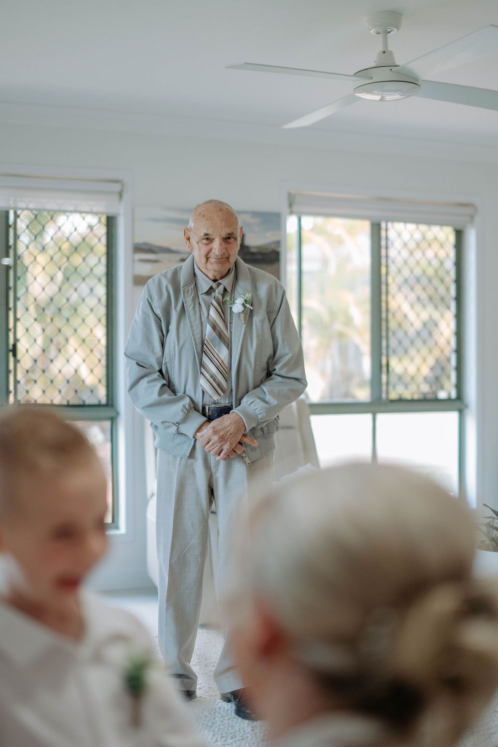 Elderly man in gray suit with striped tie stands smiling at a wedding ceremony indoors.