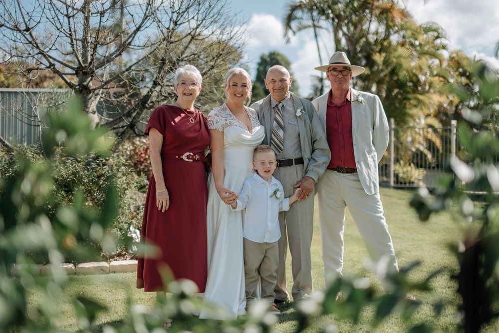 Wedding party of five posing in a garden, bride in white, guests in burgundy and grey formal attire.