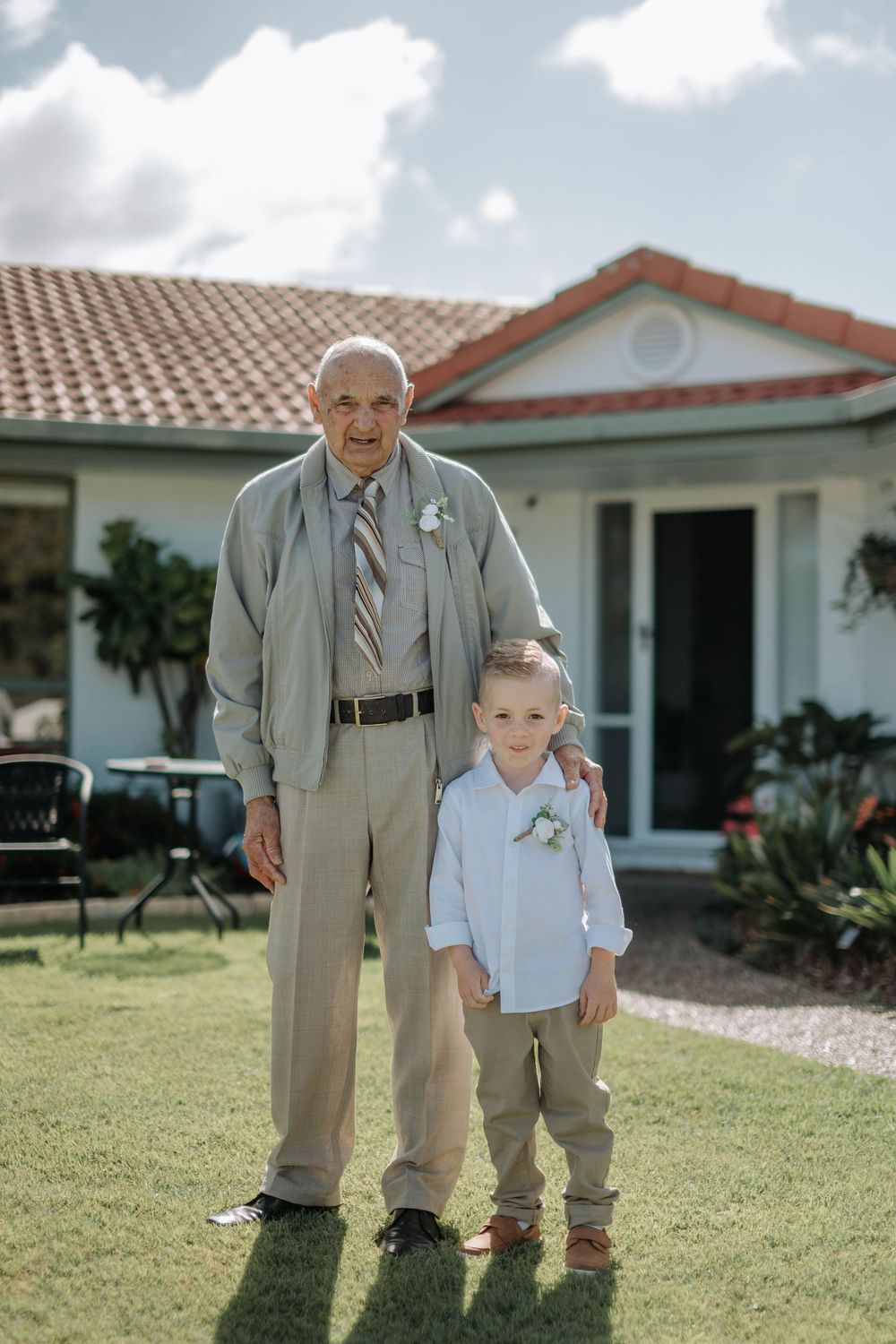 Elderly man and young boy in matching tan suits with boutonnieres pose together on lawn outside home.