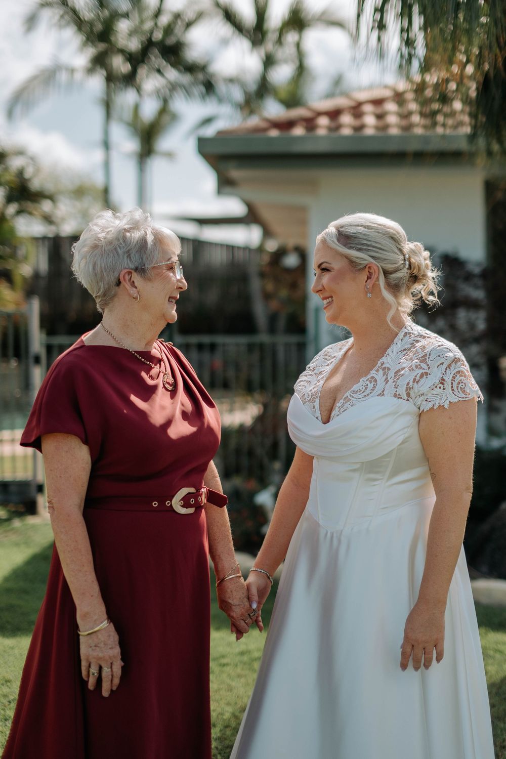 A bride in a white lace gown holds hands with an older woman in a red dress, smiling at each other outdoors.