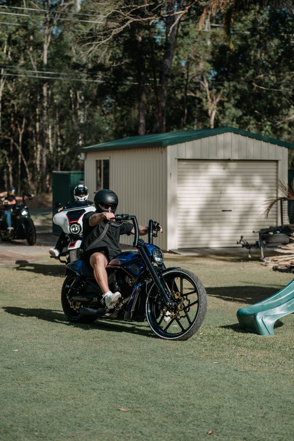 Motorcyclist in helmet riding custom chopper on grass with garage and trees in background.