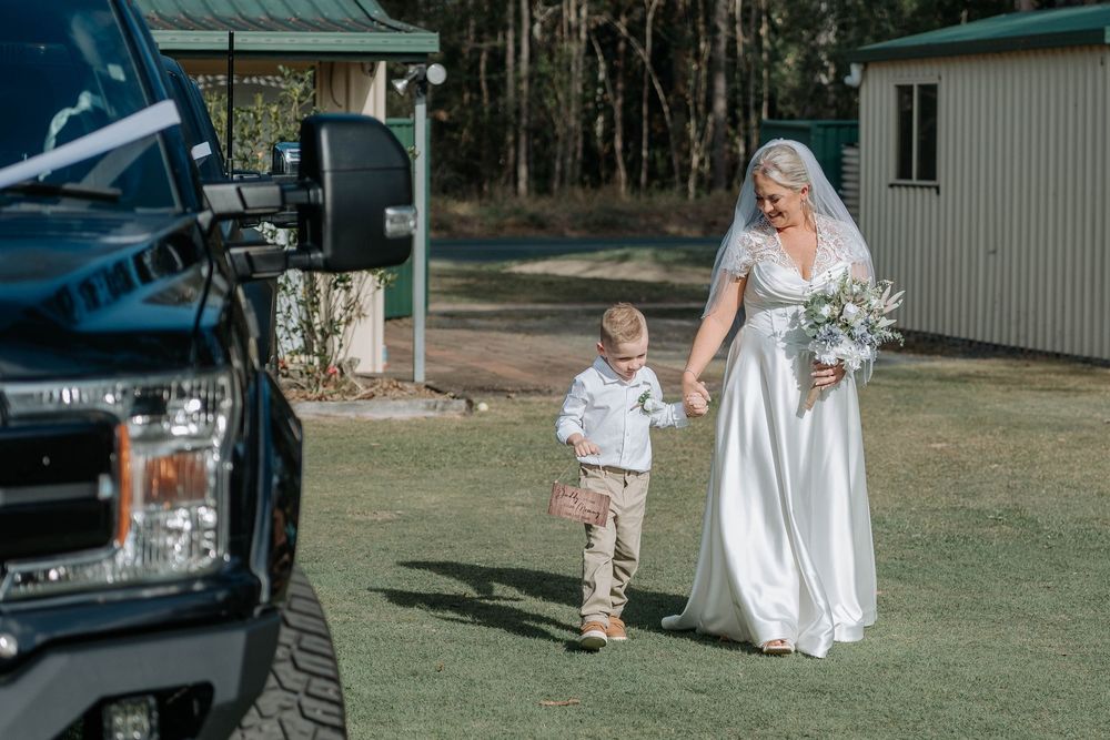 Bride in white gown holding bouquet walks with young boy on green lawn near black SUV at outdoor wedding venue.