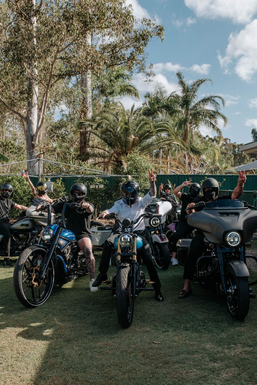 Group of motorcyclists wearing helmets posing on Harley-Davidson bikes on grass with palm trees in background.