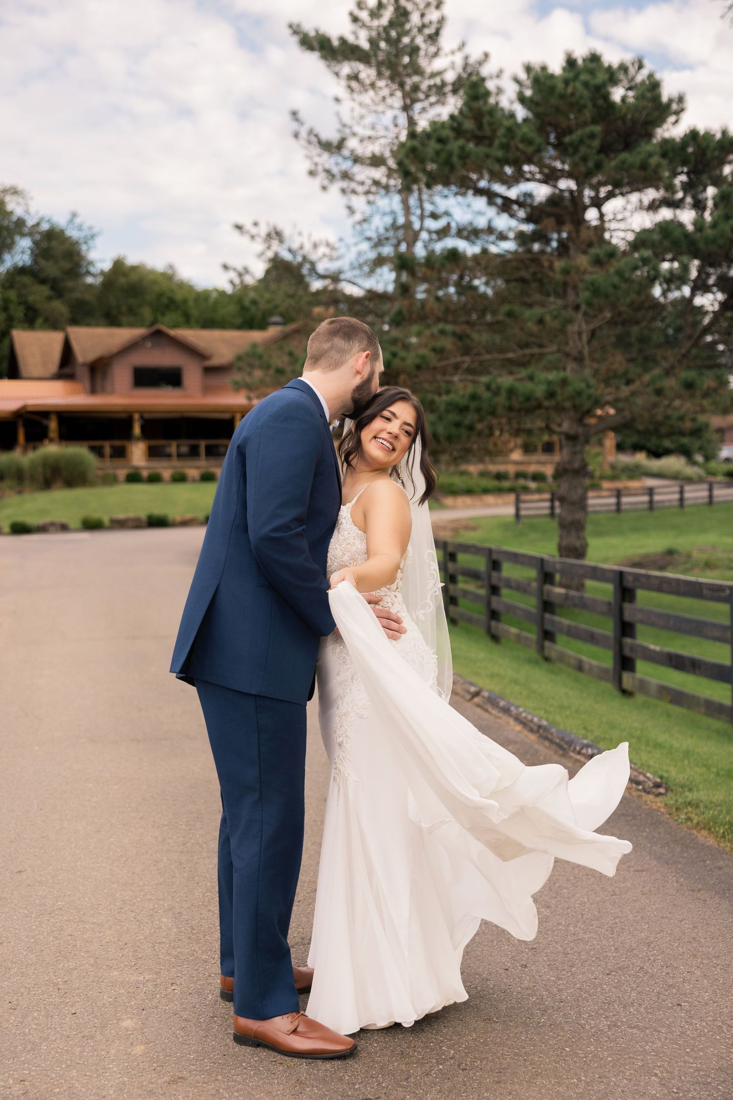Bride in white gown and groom in navy suit share a romantic embrace on a path at a rustic farm wedding venue.