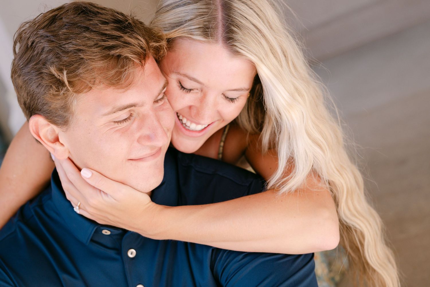 A young couple shares a tender embrace, the smiling blonde woman hugging the man from behind outdoors.