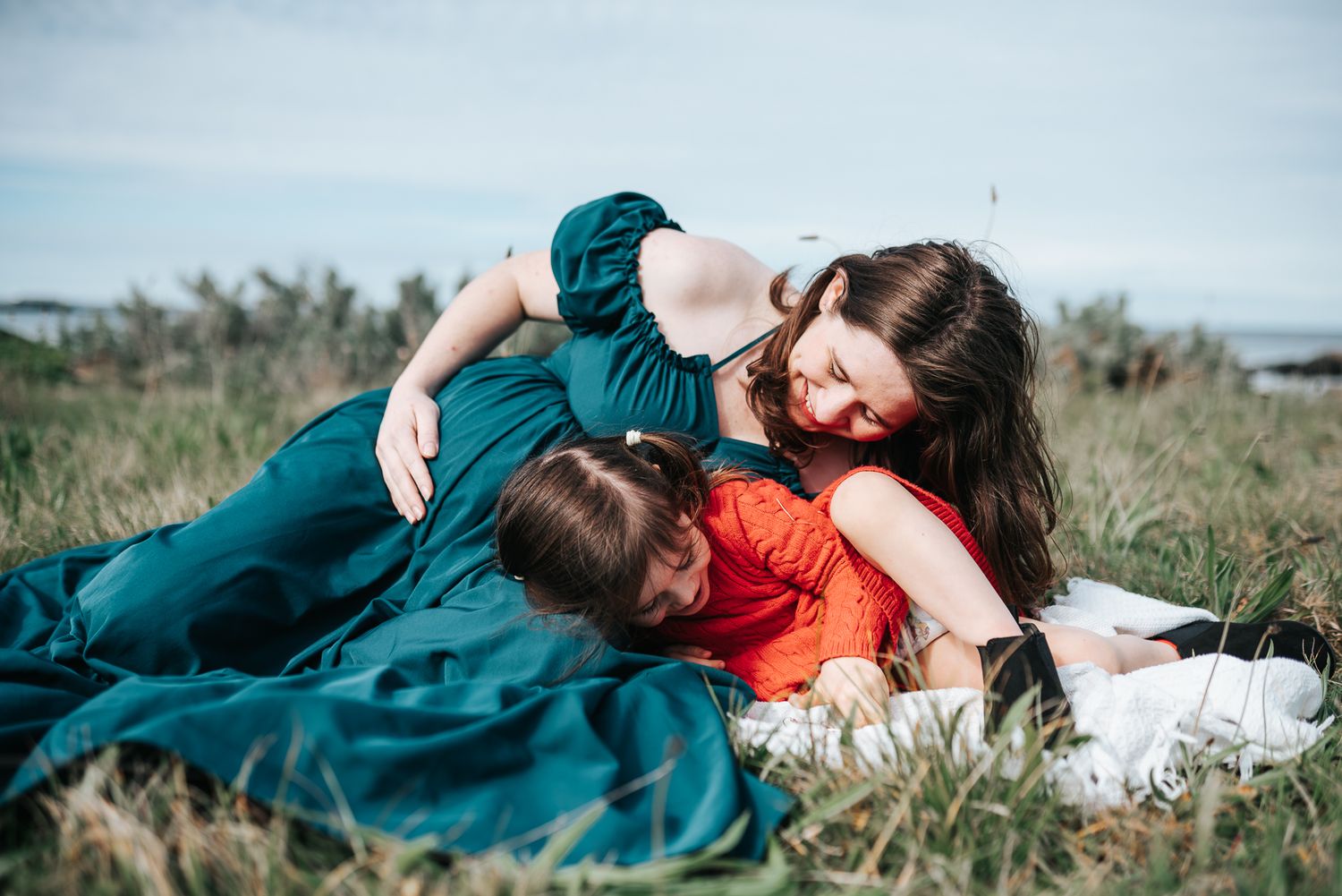 A candid photo of a pregnant woman in teal coloured dress and her daughter lying together in grass field taken by Melbourne photographer.