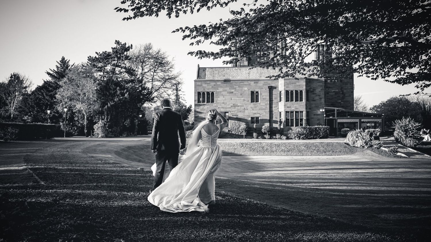 Black and white photograph of a newly married couple walking together on the lawn beside their wedding venue.