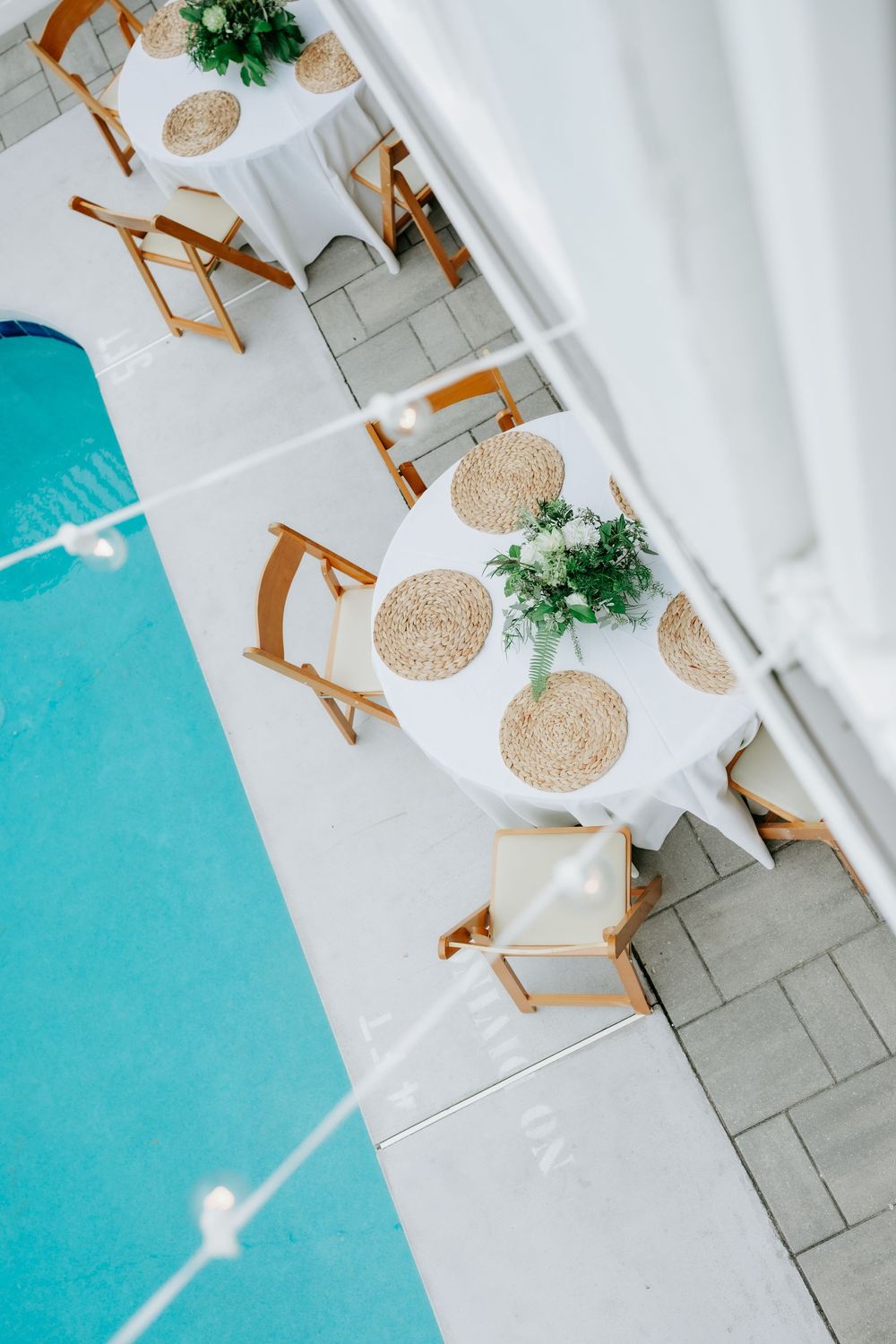 Aerial view of elegant poolside dining tables with white linens, wicker placemats, and wooden chairs beside a turquoise pool.