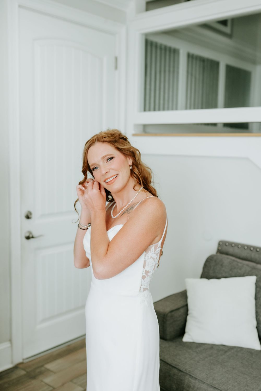 Smiling bride in white gown adjusting earring, wearing pearl necklace, standing in bright room before wedding ceremony.