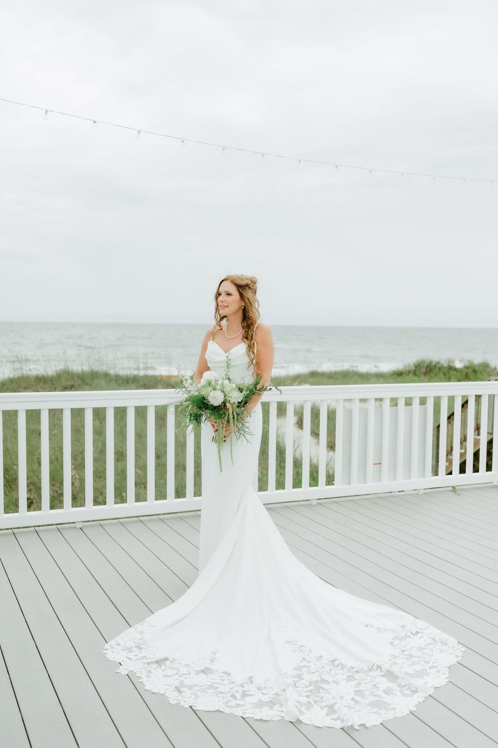 Bride in white lace mermaid gown holding bouquet on beachfront deck with ocean views.
